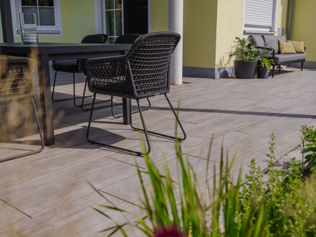 anthrazitfarbene Gartenmöbel auf Terrassenplatten in Holzoptik Calido Grano Grau vor gelber Hauswand